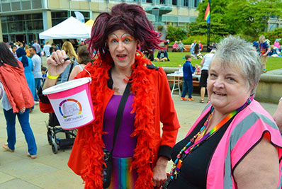 Photo of two individuals at Pinknic with the event in the background. The person on the left has an incredibly colourful outfit and is holding a pink donation bucket for LGBT Sheffield. The individual on the right has short white hair and is wearing a pink high-visibility jacket.