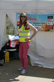 An individual in a green high vis jacket and pink trousers standing in front of a white tent holding a clipboard and a donation bucket.
